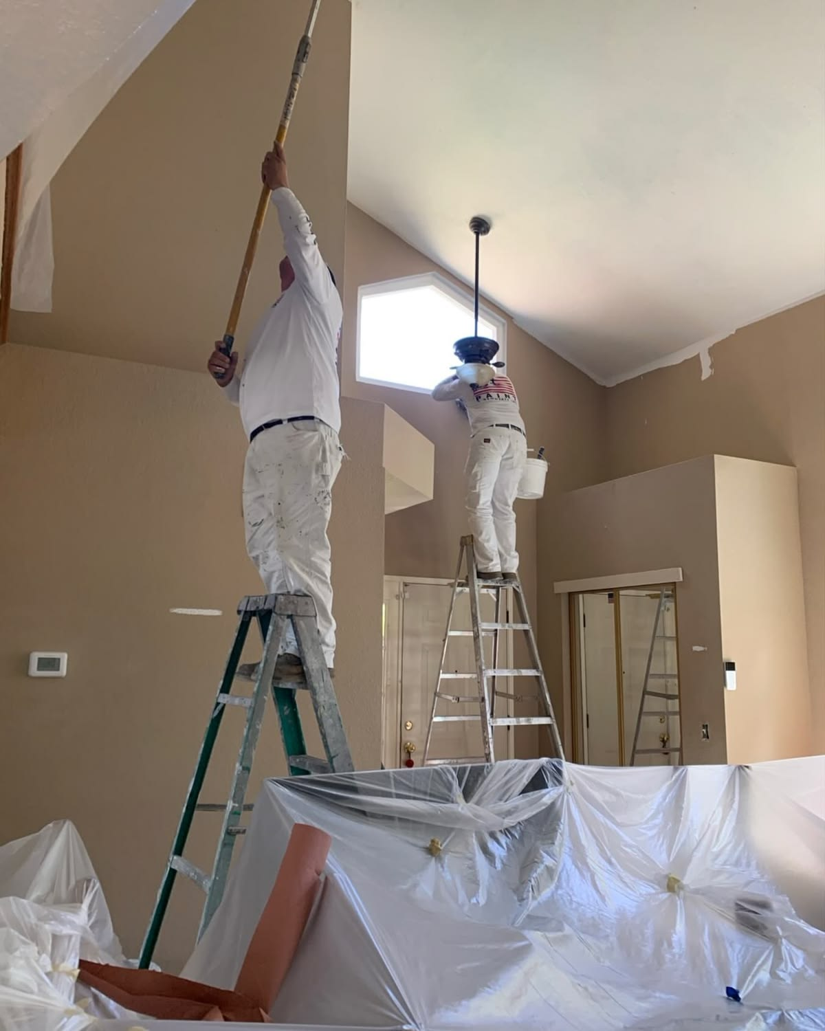 Two painters on ladders rolling a vaulted ceiling over covered furniture